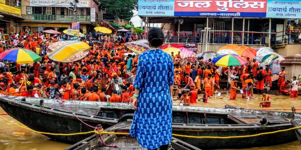 Woman dressed in blue kurta standing in a boat looking at the Kanwariyas bathing in the ganges