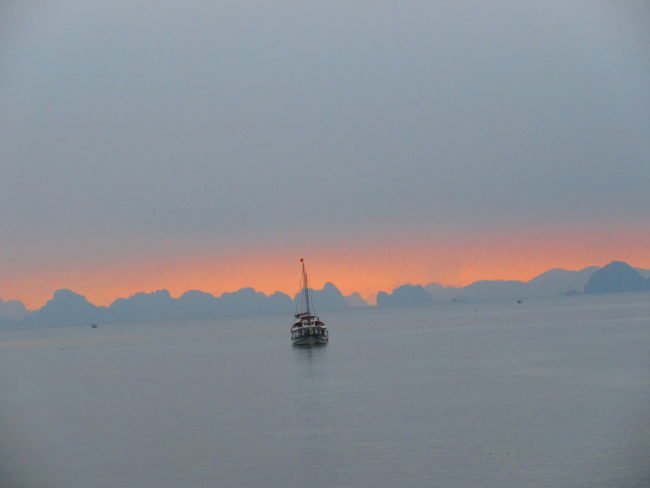 a ship sailing in the calm waters of Vietnam with the sky having an orange hue of sunset